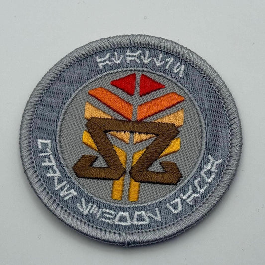 At-Attin Middle School Gamma embroidered patch, featuring vibrant orange, red, and brown geometric designs with Aurebesh text encircling the outer edge, on a light gray background.